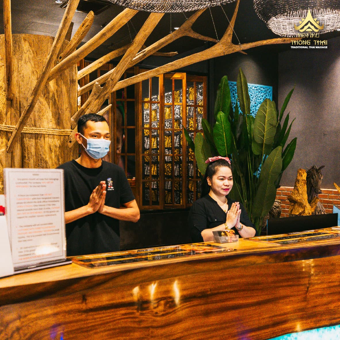 Two staff members at a wooden reception desk performing a traditional Thai wai greeting.